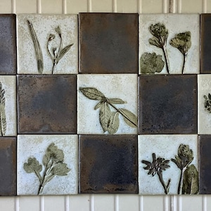 May include: A grid of fifteen square ceramic tiles. The tiles alternate between dark, mottled brown and light, speckled beige. Each beige tile features a raised botanical design in green and brown, depicting various plants. The tiles are arranged in a 3x5 pattern.