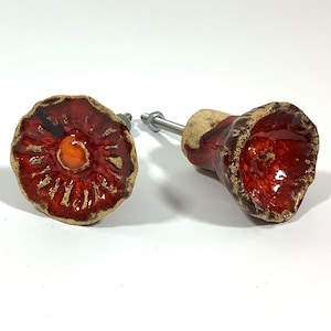 May include: Two ceramic drawer knobs in a red and brown glaze. One knob is a flat flower shape with a raised centre. The other knob is a bell shape with a rounded top.