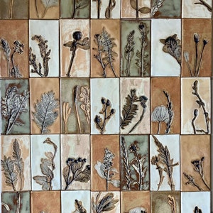 Op de afbeelding: Een wand van 36 kleine keramische tegels, elk met een ander geperst botanisch ontwerp. De tegels zijn in een rasterpatroon gerangschikt en hebben een lichtbruine achtergrond met witte voegen.