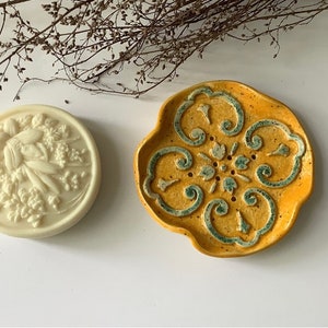 May include: A white soap bar with a floral design sits on a yellow and green ceramic soap dish. The dish has a decorative pattern and a scalloped edge.