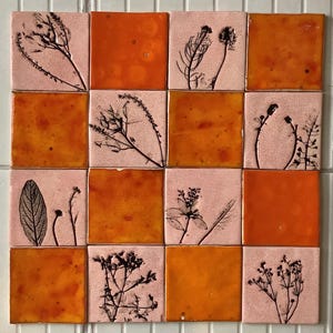 Op de afbeelding: Een set van 12 vierkante tegels met een roze achtergrond en oranje glazuur. Elke tegel heeft een geperst botanisch ontwerp in zwart.