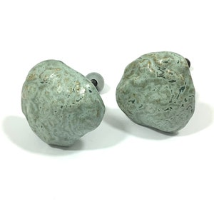 May include: A pair of ceramic cabinet knobs, each resembling a small, textured rock. The knobs are a muted green-grey with brown highlights. They are mounted on silver-coloured metal bases. The knobs would suit a kitchen or bathroom.