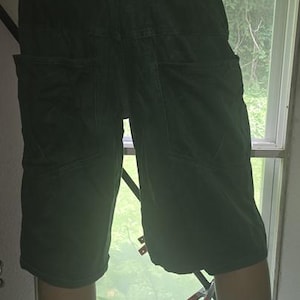 May include: Dark green denim shorts with large back pockets. The shorts are displayed on a mannequin with light skin tone legs. The shorts are knee-length and have a classic five-pocket design.