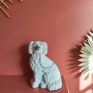 Puede incluir: Una figura de perro de cerámica blanca con collar de cadena y textura de pelaje detallada. El perro está sentado sobre una superficie rosa contra una pared rosa. Elementos decorativos: espejo de sol y hoja de palma seca.