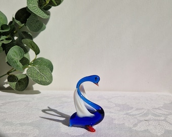 May include: A blue and white glass swan figurine with a red beak and feet. The swan is in a graceful pose with its neck curved and its head tilted.