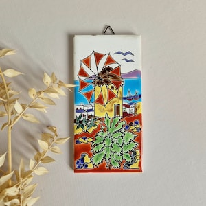 May include: A colourful ceramic tile featuring a painted image of a windmill, palm tree, and houses. The windmill has red and white blades and a yellow roof. The palm tree has green leaves and the houses are white with blue accents. The tile has a brown background and a blue sky with white birds.