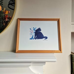 May include: A framed art print featuring a navy blue silhouette of a cat and a cluster of blue and white flowers. The artwork is displayed on a white surface, with a green vase and a mirror in the background.