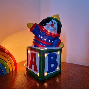 Può includere: Lampada a forma di clown in ceramica dal design colorato. Il clown è appollaiato su un blocco con le lettere "A" e "B". La lampada ha un cappello giallo, un abito rosso e blu ed è illuminata da piccole luci. Un arcobaleno di legno è sullo sfondo.