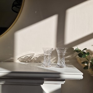 May include: Two clear glass bud vases with a cut glass design sit on a white mantelpiece. The vases are empty and the mantelpiece is illuminated by sunlight.