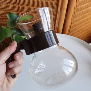 Può includere: Una caffettiera in vetro trasparente con un manico e un coperchio in plastica marrone. La caffettiera ha un corpo rotondo e un beccuccio. Il testo "Use & Care: Rinse only to clean. Do not bump or scratch. Made in U.S.A." è stampato sul fondo della caffettiera.