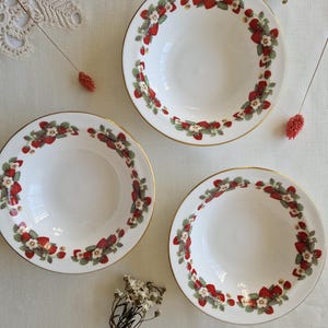 May include: Three white ceramic bowls with a gold rim and a strawberry design. The bowls are on a white surface with a lace doily and dried flowers.