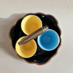 May include: A black ceramic serving tray with three small cups. The cups are yellow, blue, and yellow. A metal handle is attached to the tray.