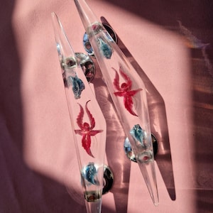 May include: Two clear, pointed cabinet pulls with embedded red and blue decorative elements. The pulls are mounted on silver-colored circular bases. The background is a solid pink color.