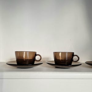 以下が含まれることがあります： 茶碗と受け皿が3つ。茶碗は丸みを帯びた形をしていて、小さな取っ手がついています。受け皿は丸く、縁が少し盛り上がっています。茶碗と受け皿は茶色いガラス製です。