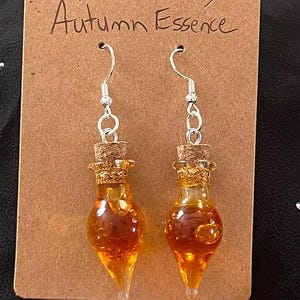 May include: A pair of dangle earrings featuring small glass bottles with cork stoppers filled with a golden liquid. The earrings are silver and have a hook closure. The card behind the earrings says "Autumn Essence".