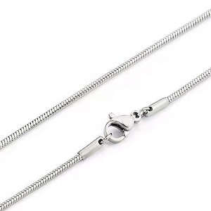 Puede incluir: Un collar de cadena de serpiente de color plateado con cierre de mosquetón. La cadena tiene una textura suave y redondeada y se muestra sobre un fondo blanco. El collar es una joya sencilla y elegante.
