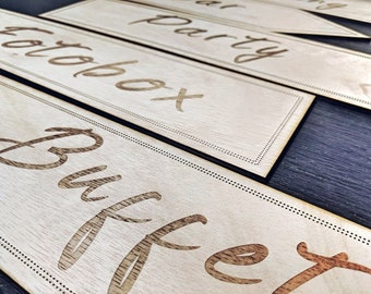May include: Wooden signs with laser-etched lettering for events. The signs read "Fotobox", "Party", and "Buffet" in a decorative font. Each sign has a dotted border and is made of light-colored wood.