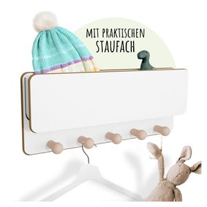 Könnte beinhalten: Ein weißer Wandorganizer mit einem Regal und fünf Holzknöpfen. Eine hellblau-weiß gestrickte Mütze und eine kleine grüne Dinosaurierfigur liegen auf dem Regal. Der Organizer enthält auch einen weißen Kleiderbügel und ein Stofftier.