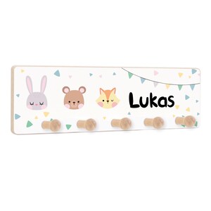 Könnte beinhalten: Ein weißer Holz-Kleiderhaken mit fünf Haken. Der Kleiderhaken hat ein farbenfrohes Design mit einem Hasen, einem Bären und einem Fuchs. Der Name "Lukas" ist in schwarzen Buchstaben auf dem Kleiderhaken geschrieben.