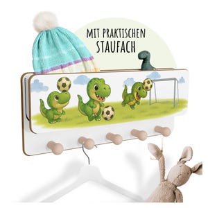 Könnte beinhalten: Ein weißer Wandorganizer mit einem verspielten Dinosaurier-Fußball-Thema. Der Organizer verfügt über ein Regal, Holzknöpfe und einen Kleiderbügel. Oben steht der Text "MIT PRAKTISCHEN STAUFACH". Eine blau-weiß gestrickte Mütze und ein Stofftier befinden sich auf dem Organizer.