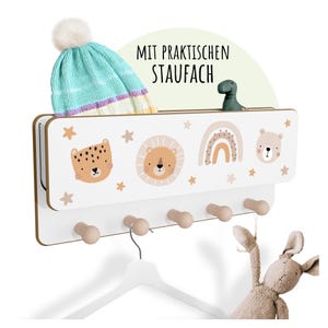 Könnte beinhalten: Ein weißer und holzfarbener Wandorganizer mit Tierillustrationen und Holzknöpfen. Der Organizer hat ein Regal mit einer blau-weißen Strickmütze und einer kleinen grünen Dinosaurierfigur. Der Text "MIT PRAKTISCHEN STAUFACH" ist sichtbar.
