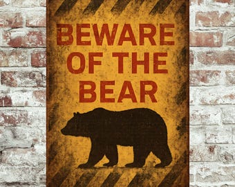 May include: A rectangular metal sign with a distressed yellow background and the words "BEWARE OF THE BEAR" in red. A black silhouette of a bear is below the text. Diagonal black and yellow stripes are at the top and bottom.