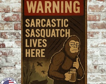 Op de afbeelding: Een rechthoekig metalen bord met een bruine achtergrond en een rode banner met de tekst "WARNING". Het bord toont de tekst "SARCASTIC SASQUATCH LIVES HERE" en een illustratie van een Sasquatch met een pot. Linksonder staat een "MADE IN USA" embleem.