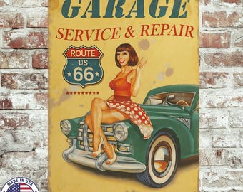 May include: Vintage metal sign with a distressed finish. The sign features the words "GARAGE SERVICE & REPAIR" above a Route 66 emblem. A pin-up girl in a red dress sits on a green classic car. The sign has a "Made in USA" emblem.