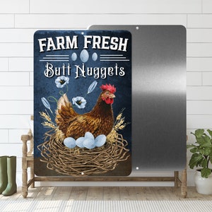 May include: A blue and white metal sign with the text "FARM FRESH Butt Nuggets" and a cartoon image of a brown hen sitting on a nest of eggs.