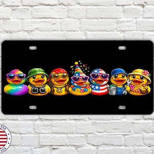 May include: Black license plate featuring seven cartoon ducks, each with unique accessories like sunglasses, hats, and outfits. The ducks are arranged in a row, with vibrant colors and detailed designs. The plate has a "Made in USA" emblem.