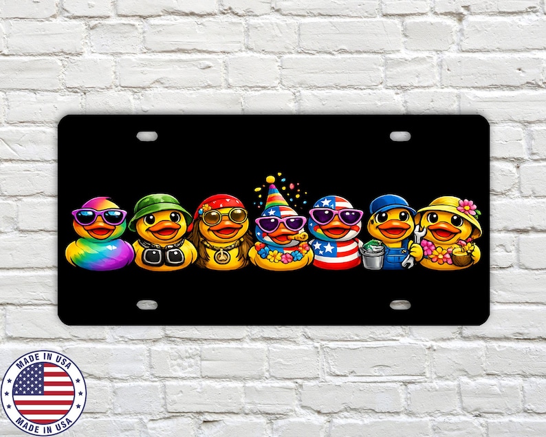 May include: Black license plate featuring seven cartoon ducks, each with unique accessories like sunglasses, hats, and outfits. The ducks are arranged in a row, with vibrant colors and detailed designs. The plate has a "Made in USA" emblem.