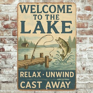 Op de afbeelding: Vintage metalen bord met de tekst "WELCOME TO THE LAKE" in blauw. Het bord toont een visscène met een pier, een springende vis en de zinnen "RELAX • UNWIND" en "CAST AWAY".