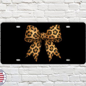 May include: Black license plate featuring a large leopard print bow. The bow is in shades of brown and tan with black spots. The license plate has four mounting holes.