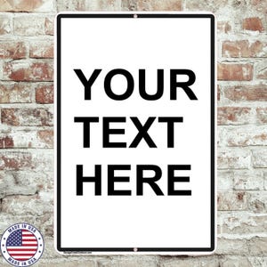 May include: A rectangular white sign with black text that reads "YOUR TEXT HERE." The sign has a black border and is mounted on a brick wall. A small "Made in USA" sticker with an American flag is in the lower left corner.