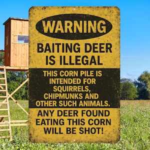 May include: A distressed yellow and black metal sign with the text "WARNING BAITING DEER IS ILLEGAL." The sign also states the corn pile is for squirrels, chipmunks, and other animals. A wooden hunting blind is in the background.