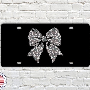 May include: Black license plate featuring a large bow design in shades of gray and white with a camouflage pattern. The license plate has four mounting holes and a glossy finish. The words "MADE IN USA" are printed on a circular sticker in the lower left corner.
