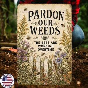 May include: A rectangular metal garden sign with a cream background featuring the black text PARDON OUR WEEDS THE BEES ARE WORKING OVERTIME. The design includes illustrations of a white picket fence, yellow and purple wildflowers, and bees.