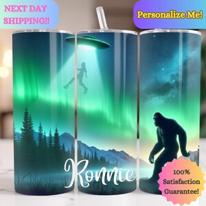 Op de afbeelding: Gepersonaliseerde roestvrijstalen tumbler met een groen en blauw aurora borealis-ontwerp. De tumbler heeft een silhouet van een persoon die door een UFO wordt ontvoerd en een Bigfoot die door het bos loopt. De tumbler is gelabeld met "Ronnie".