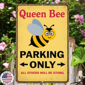 May include: A yellow metal sign featuring a cartoon bee and the words "Queen Bee" in red. The sign reads "PARKING ONLY" with arrows and "ALL OTHERS WILL BE STUNG" in black. A "Made in USA" emblem is in the lower left corner.