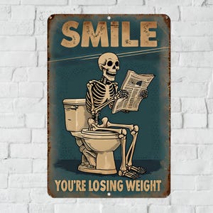 May include: A vintage-style metal sign with a humorous illustration. It features a skeleton sitting on a toilet, reading a newspaper, with the words "SMILE" and "YOU'RE LOSING WEIGHT" in bold text. The sign has a distressed, aged appearance.