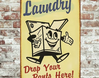 May include: A vintage-style laundry sign with a yellow background and a cartoon box character. The sign reads "Laundry" in blue script and "Drop Your Pants Here!" in red. The sign is rectangular and has a distressed, aged appearance.