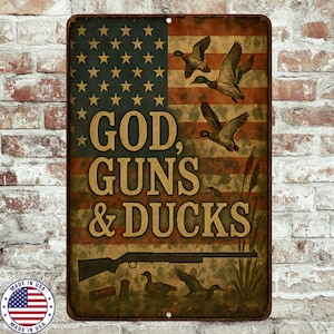 Op de afbeelding: Rechthoekig metalen bord met een Amerikaans vlagontwerp en de tekst "GOD, GUNS & DUCKS". Eenden in vlucht en een geweer zijn afgebeeld. Het bord heeft een verweerde, vintage look, met een "Made in USA" embleem.