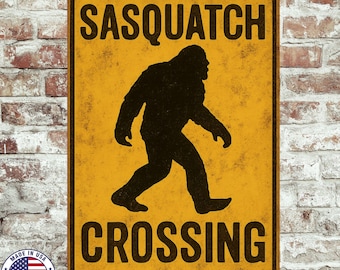 May include: A rectangular metal sign with a yellow background and black text and silhouette. The text reads "SASQUATCH CROSSING". The silhouette depicts a large, hairy creature walking. The sign has a distressed, vintage look.