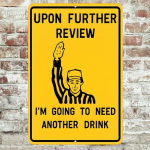 May include: A yellow metal sign with black text and a black and white illustration of a referee. The sign reads "UPON FURTHER REVIEW I'M GOING TO NEED ANOTHER DRINK."