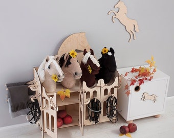 May include: Wooden toy horse stable with four plush horse heads, a brown, white, black, and brown and white horse. The stable has shelves and hooks for storage. A white cabinet with a horse silhouette is in the background.
