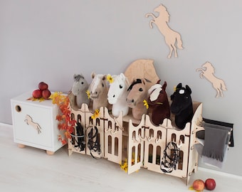 Può includere: Set da gioco di stalla per cavalli in legno con sei cavalli di peluche, ognuno con un colore e un design diverso. La stalla ha una finitura bianca dipinta e presenta una recinzione decorativa e un tetto. I cavalli sono disposti in fila, rivolti in avanti.