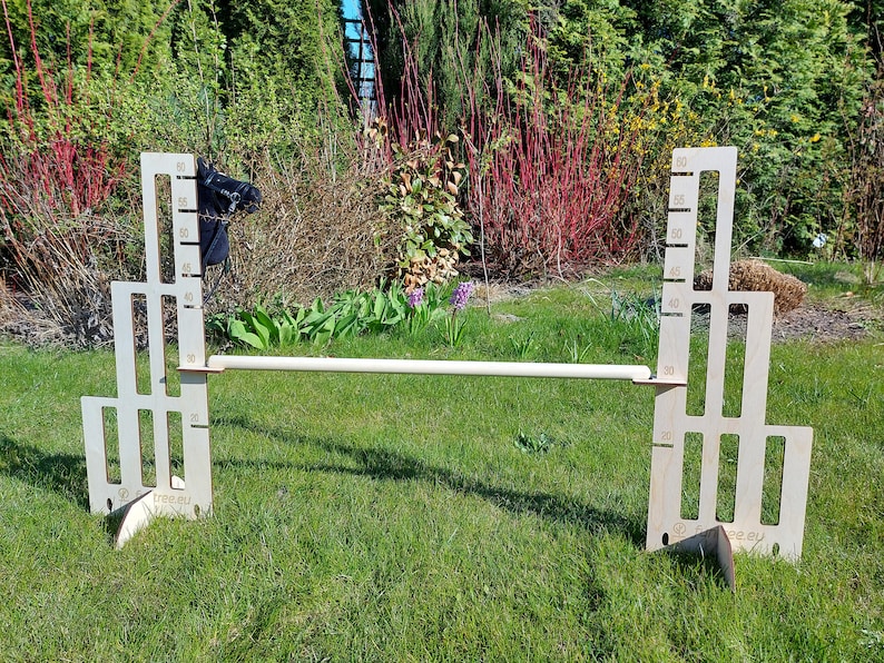 Puede incluir: Un obst&aacute;culo de salto de madera de altura ajustable con una cabeza de caballo negra en el lado izquierdo y una regla a cada lado. El obst&aacute;culo est&aacute; instalado en un c&eacute;sped verde.