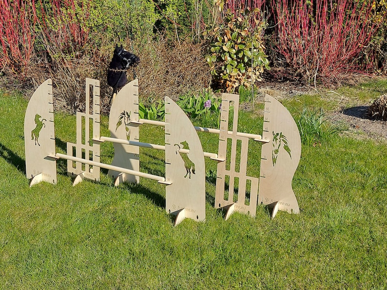 Puede incluir: Tres obst&aacute;culos de salto para caballos de madera con alturas ajustables. Cada obst&aacute;culo presenta una silueta de cabeza de caballo y una regla para medir la altura. Los obst&aacute;culos est&aacute;n instalados en una superficie cubierta de hierba.