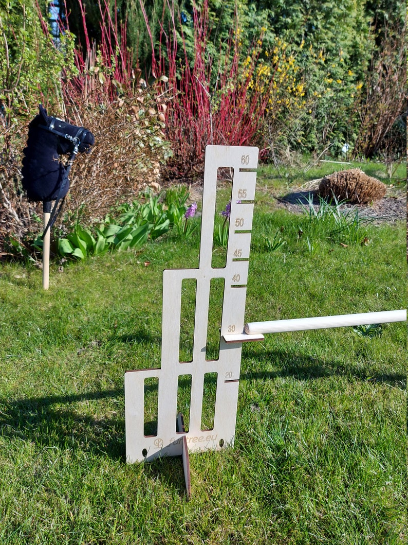 Puede incluir: Un obst&aacute;culo de salto de madera para caballos, con una altura ajustable de 50 a 150 cm. El obst&aacute;culo est&aacute; hecho de madera contrachapada y tiene un acabado blanco. El obst&aacute;culo se muestra en un campo de hierba con un juguete de cabeza de caballo negro en el fondo.