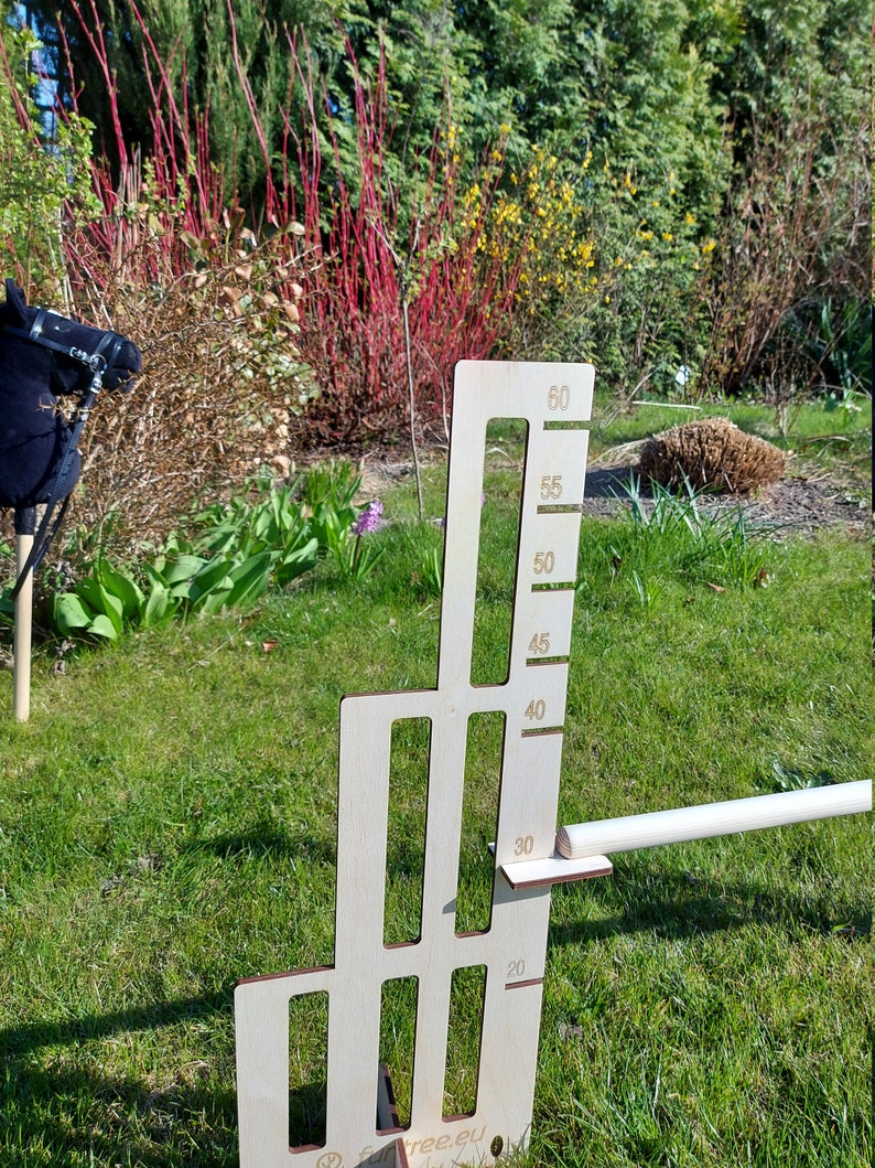 Puede incluir: Un gr&aacute;fico de altura de madera con fondo blanco y n&uacute;meros negros. El gr&aacute;fico est&aacute; marcado en incrementos de 5 cent&iacute;metros, de 20 a 60 cent&iacute;metros. El gr&aacute;fico est&aacute; de pie sobre una superficie de hierba verde.
